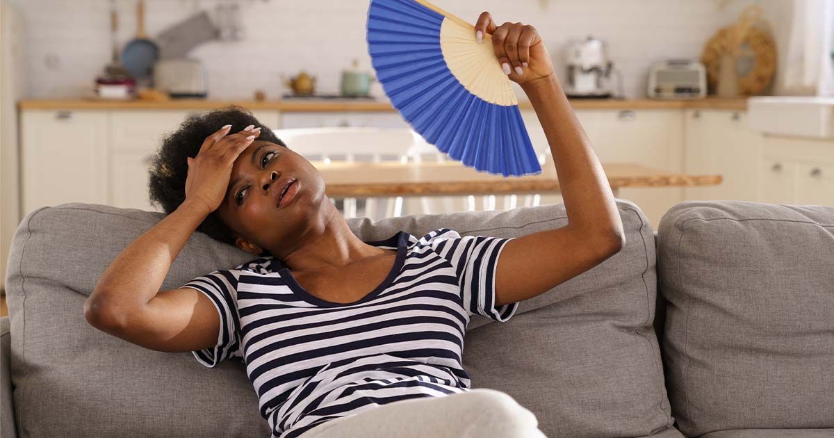 Woman sitting on couch with a fan, looking tired and overheated.